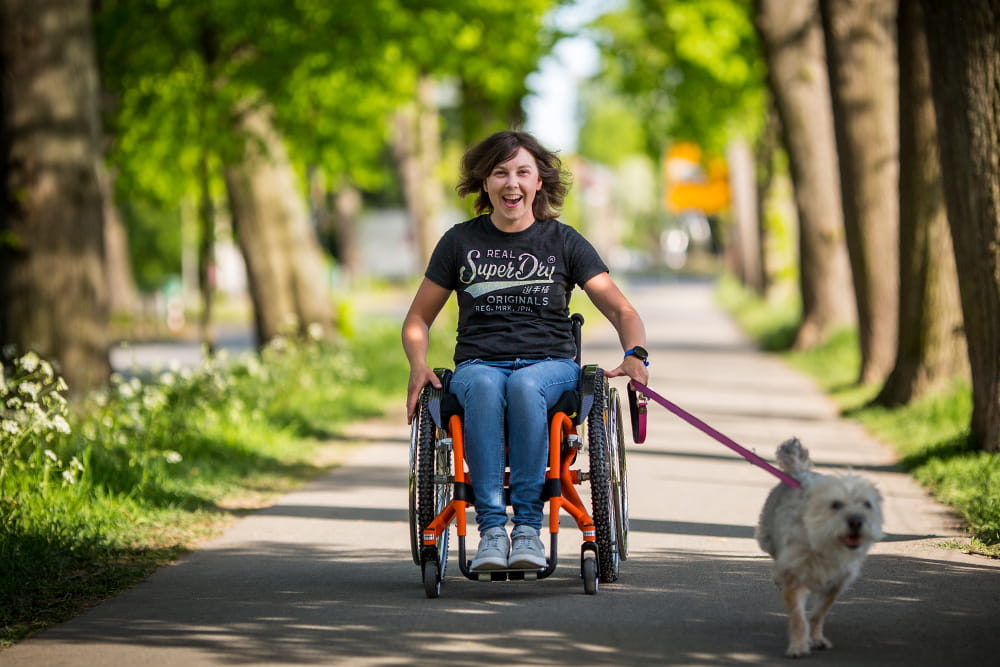 Mädchen im Rollstuhl fährt sehr schnell einen Weg entlang und ihr Hund rennt daneben her.