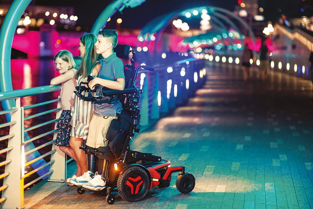 Eine Junge im Stehrollstuhl schaut mit Freunden über das Geländer auf das Wasser.