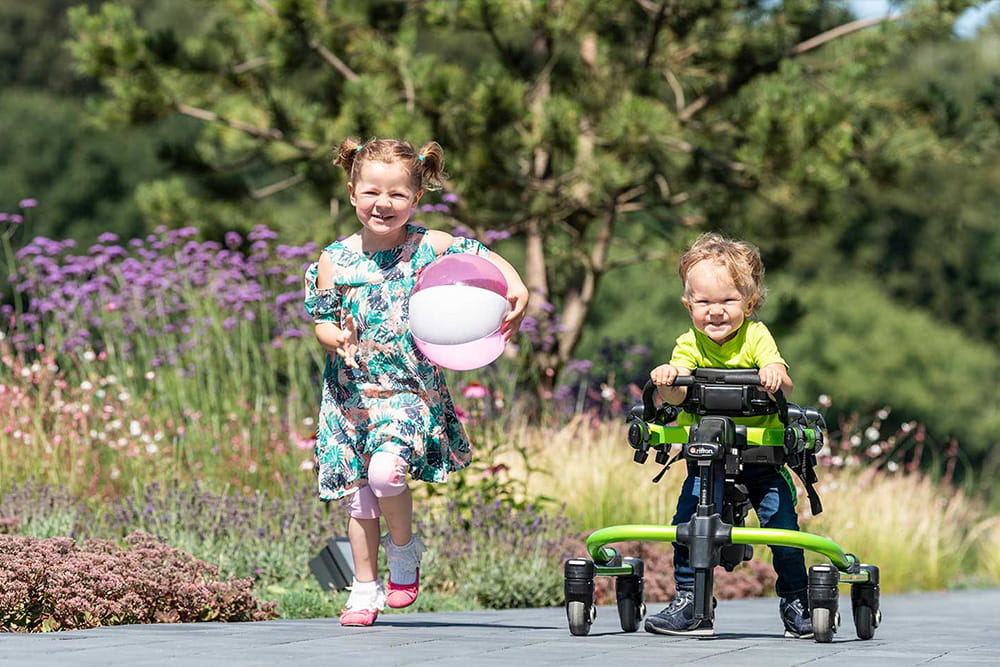 Zwei Kinder laufen im Park auf dem Weg entland. Ein Kind läuft dabei mithilfe eines Gehtrainers.
