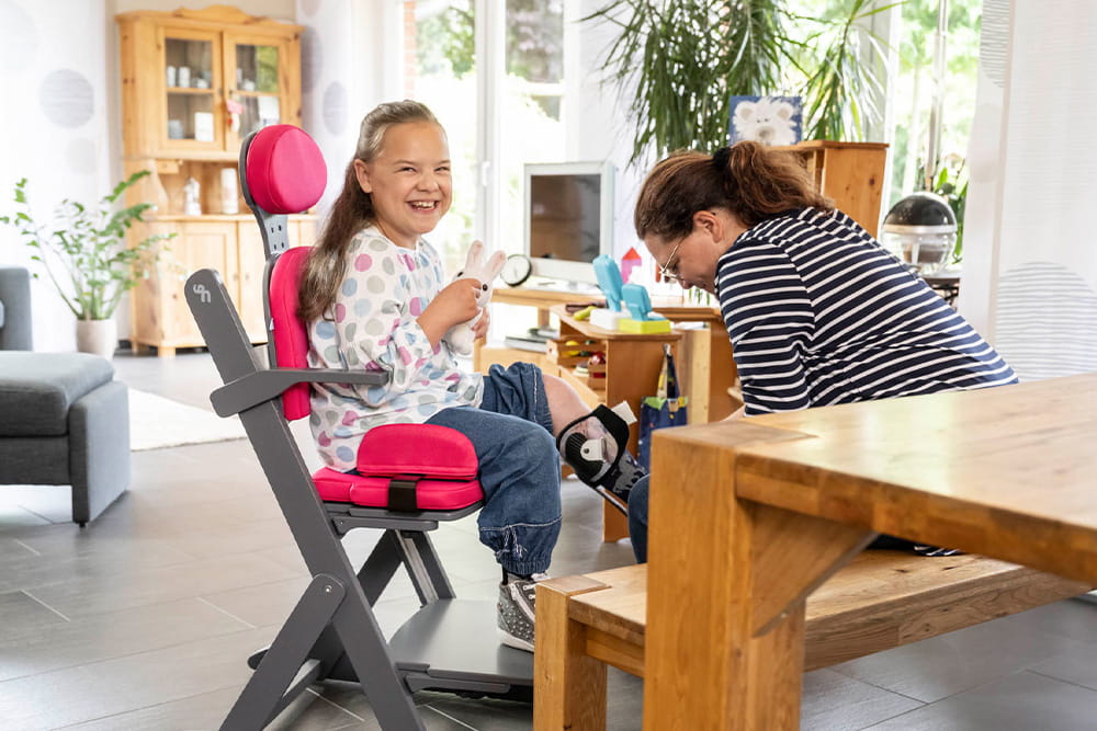 Ein Mädchen sitzt im Therapiestuhl und hält ihren Plüschhasen fest. DIe Mutter legt ihr nebenbei eine Beinorrthese an.