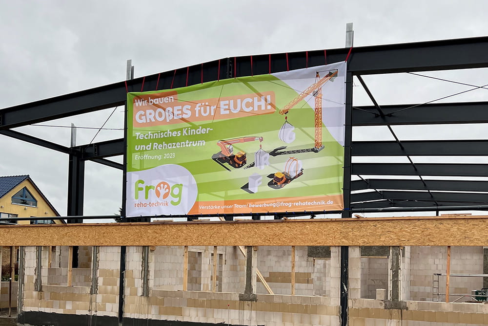 Ein Banner hängt auf der Baustelle der FROG. Das Banner kündigt das Technische Kinder- und Rehazentrum an.
