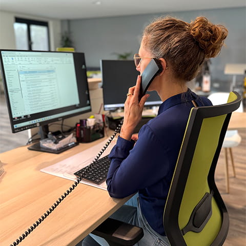 Eine Frau sitzt am Schreibtisch, schaut auf einen Monitor und hat ein Telefon in der Hand.